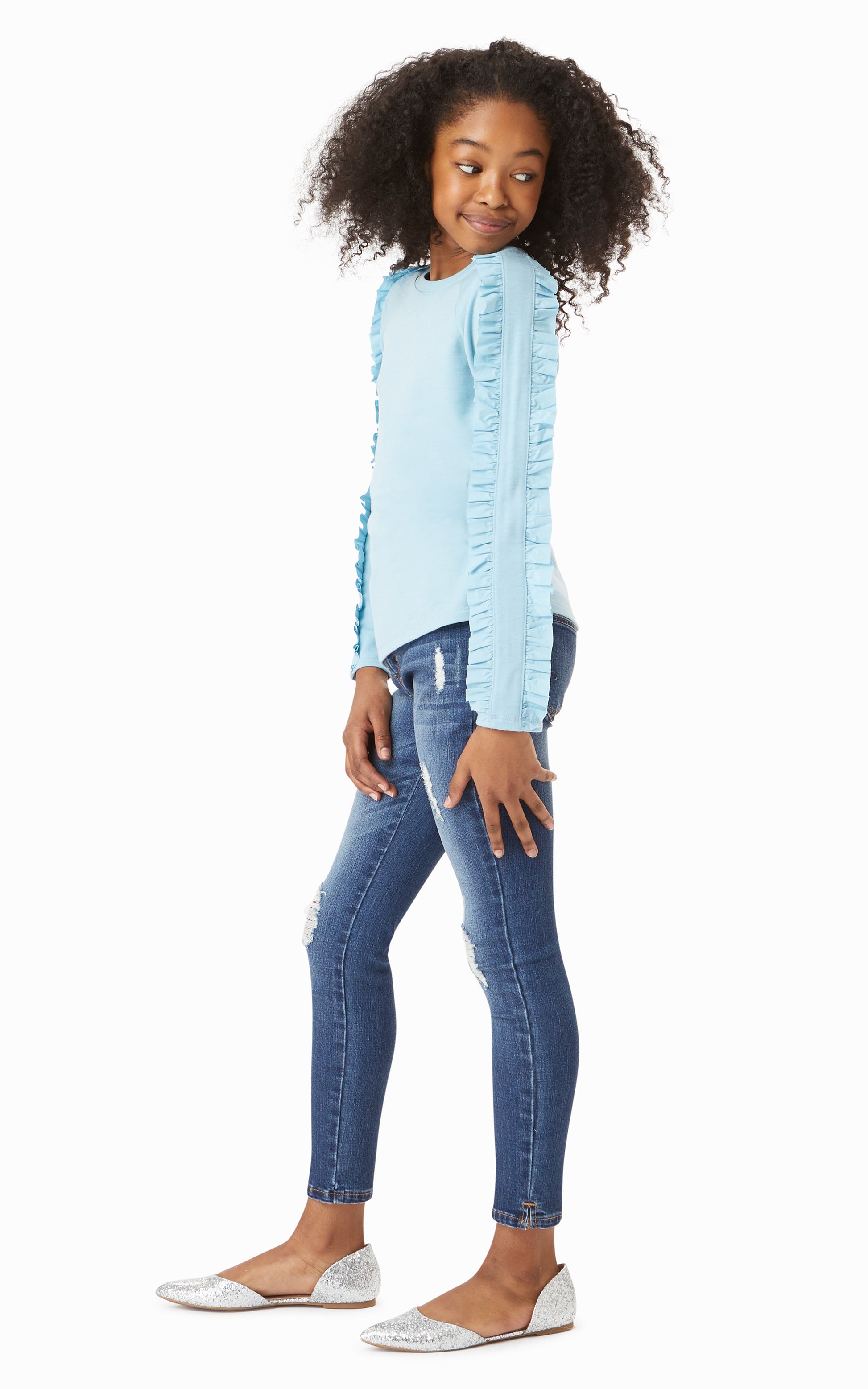 Side view of girl wearing blue long-sleeved top with ruffle trim down arms with ripped denim jeans. 