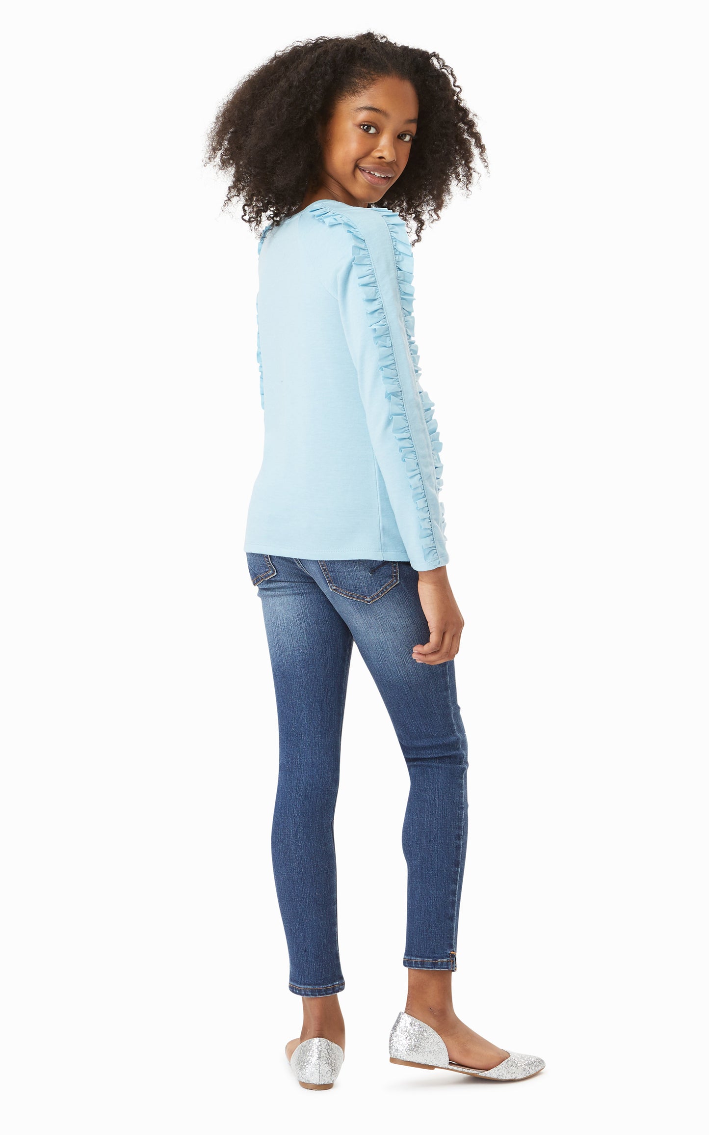 Back view of girl wearing blue long-sleeved top with ruffle trim down arms with ripped denim jeans. 