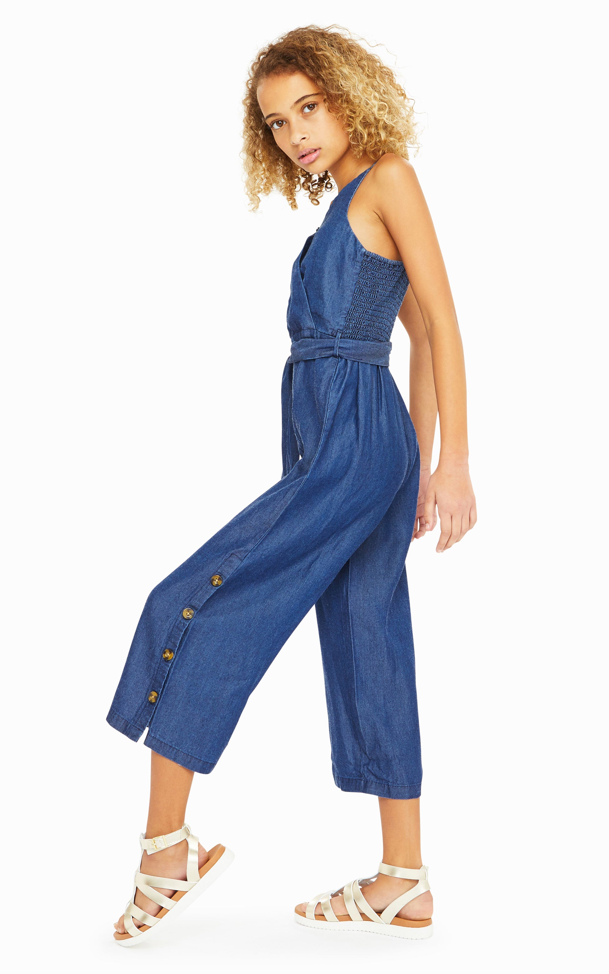 Side view of girl wearing denim-colored sleeveless jumpsuit with belted waist and leopard print button accents on legs.