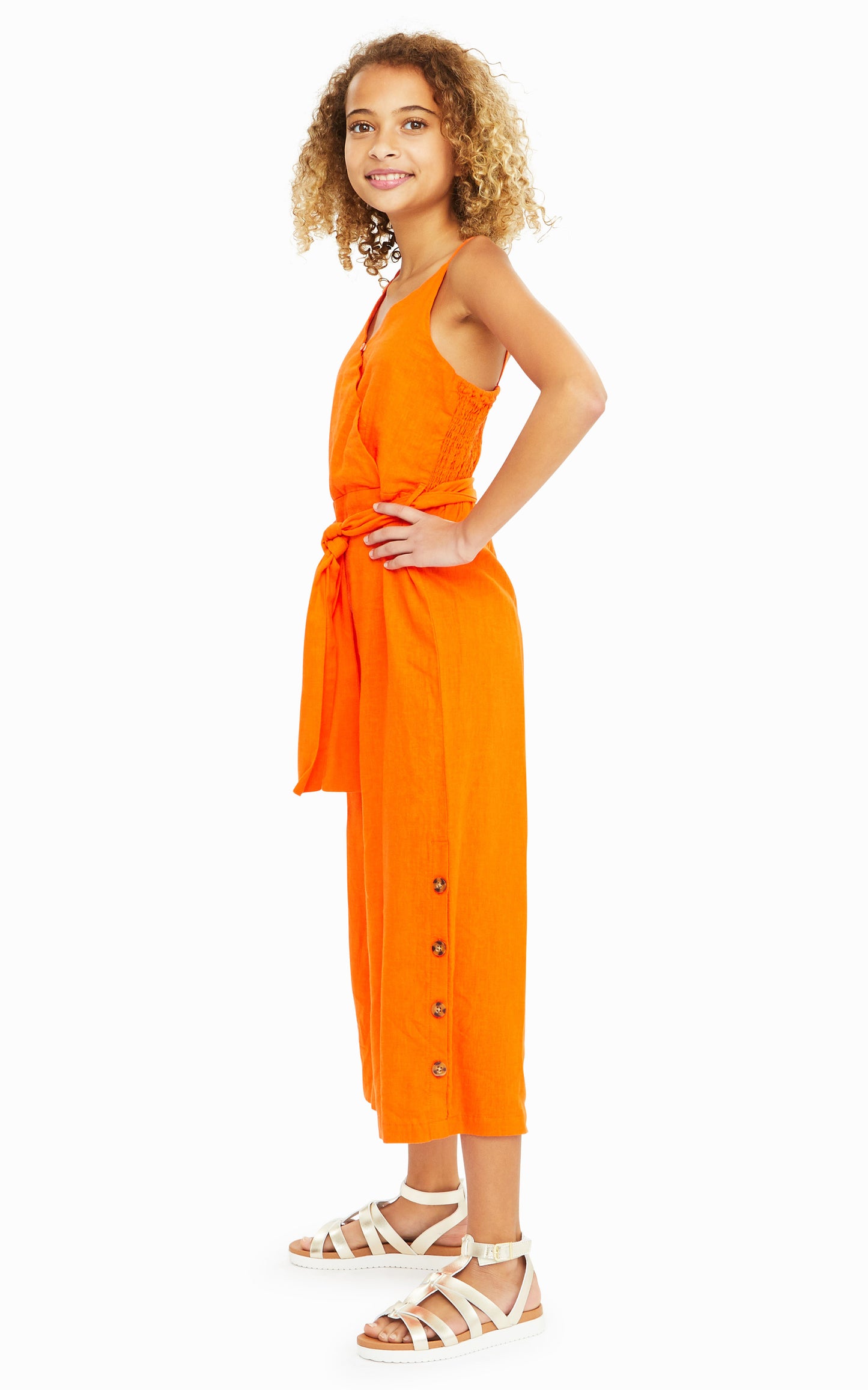 Side view of girl wearing orange sleeveless jumpsuit with belted waist and leopard print button accents on legs.