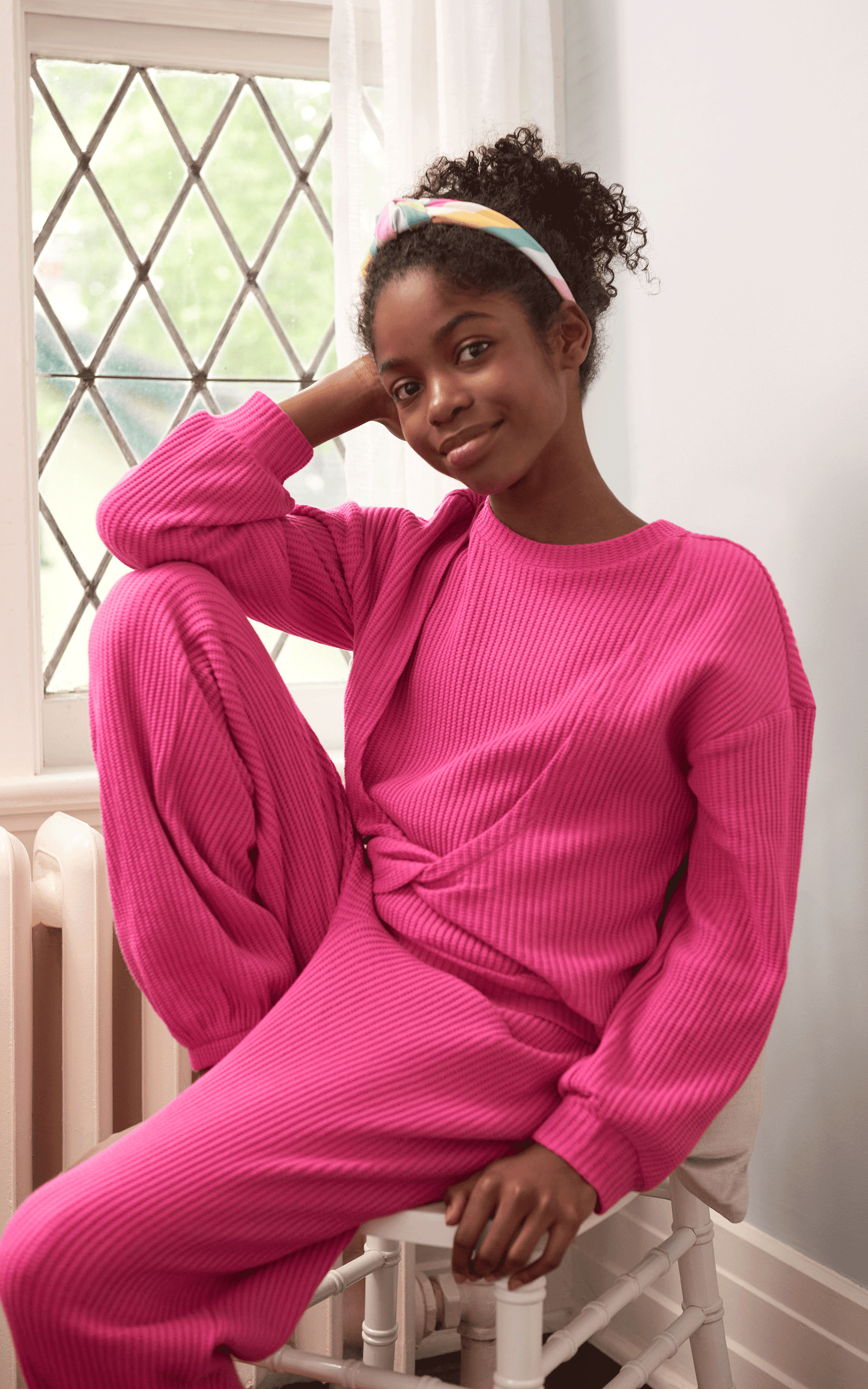 GIRL SITTING ON RADIATOR BY A WINDOW WEARING MAGENTA WAFFLE KNIT SWEATER WITH KNOT TWIST IN FRONT AND MATCHING SWEATPANTS