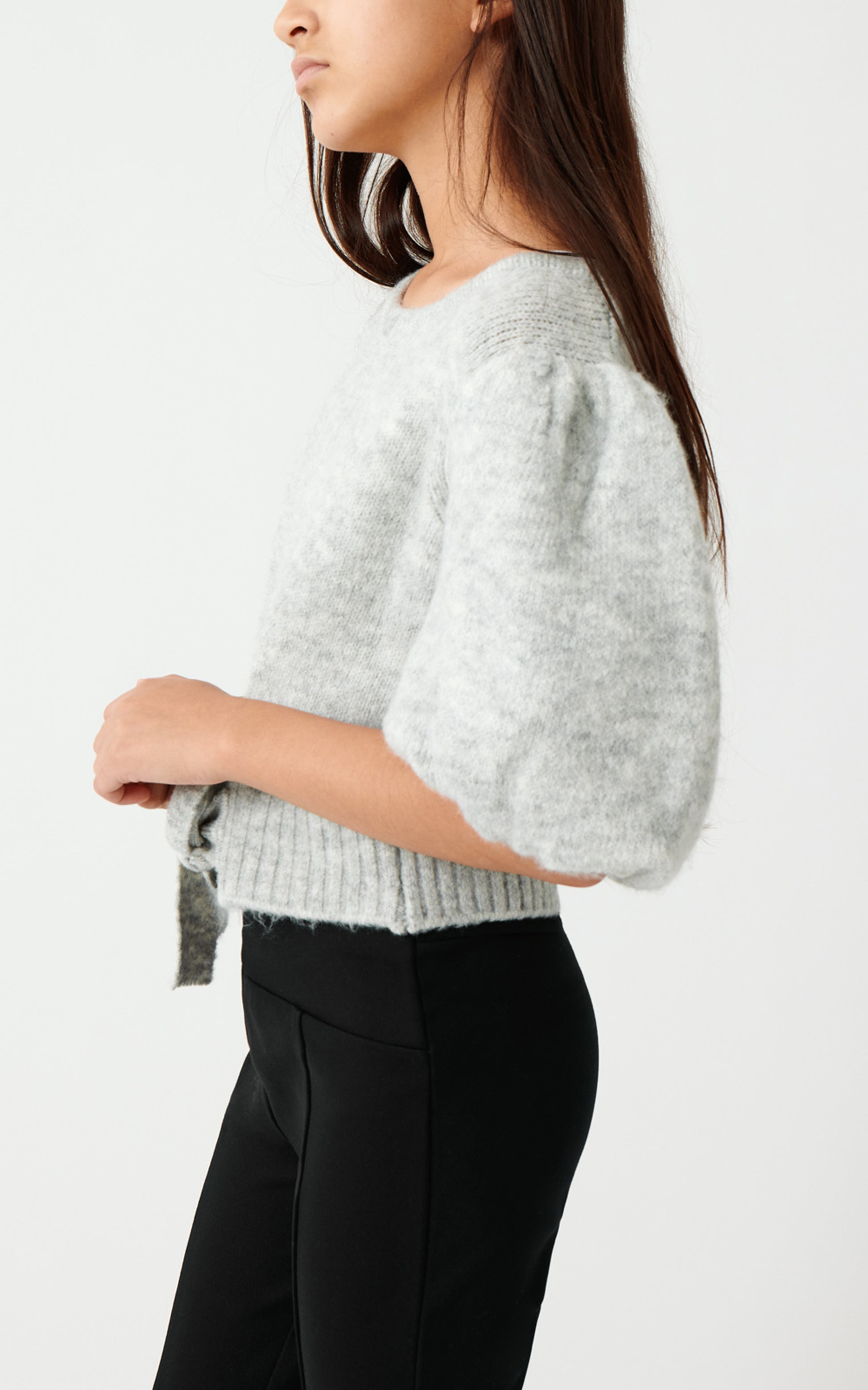 SIDE VIEW OF A GIRL WEARING AN OVERSIZED CROPPED LIGHT GREY SWEATER WITH VERY BIG PUFF SLEEVES AND BLACK PANTS